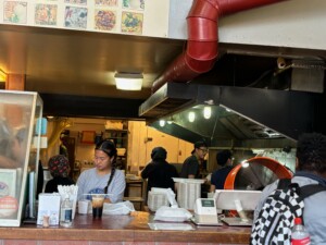 A photo of the counter of ThaiYada Restaurant. A young Asian woman with a braid stands at the counter, which is covered in Thai food and drinks ready for delivery. Behind her, the kitchen is bustling with workers.